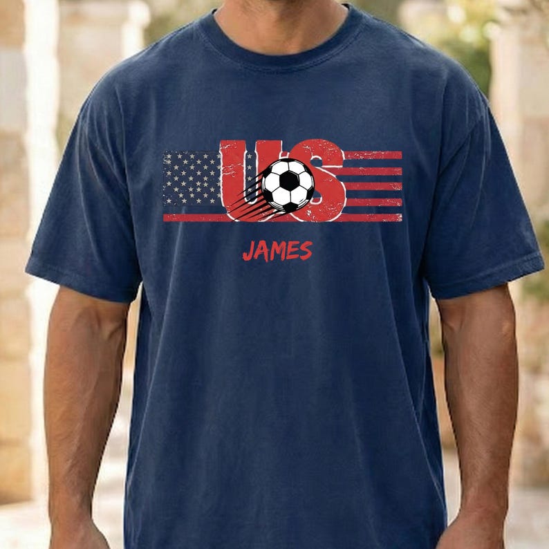 May include: A navy blue short-sleeve t-shirt featuring a distressed graphic of the American flag integrated with the letters US and a soccer ball. The name JAMES is printed in red below the graphic in a bold, sans-serif font style.