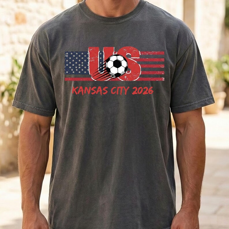 May include: A charcoal grey short-sleeve t-shirt featuring a graphic of the American flag integrated with the letters US and a soccer ball. The text KANSAS CITY 2026 is printed in red below the graphic. The design has a distressed, weathered look.