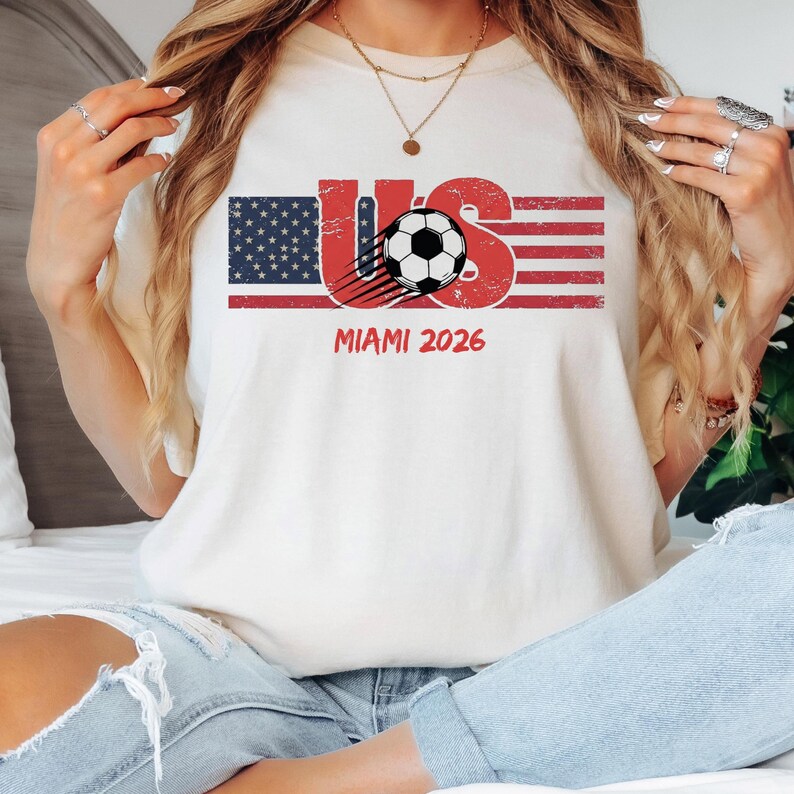 May include: A cream-colored t-shirt featuring a graphic with the text US, a stylized American flag, and a soccer ball with motion lines. Below the graphic, the text MIAMI 2026 is printed in red. The design has a distressed, weathered texture.