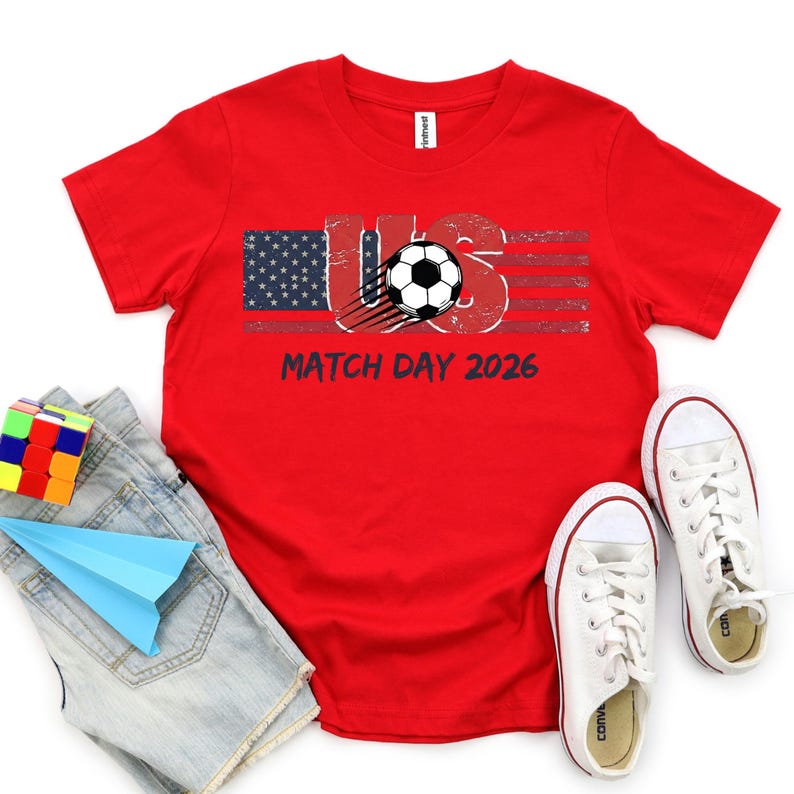 May include: A bright red short-sleeved t-shirt featuring a graphic of the United States flag integrated into the letters US, a soccer ball with motion lines, and the text MATCH DAY 2026 printed in dark blue below the central graphic design.