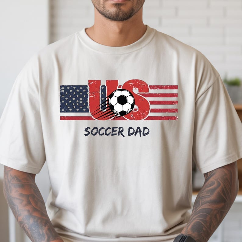 May include: A beige short-sleeved t-shirt featuring a graphic design with the text US, where the U is styled like the American flag and the S is red. A black and white soccer ball with motion lines sits in the center, with SOCCER DAD below.