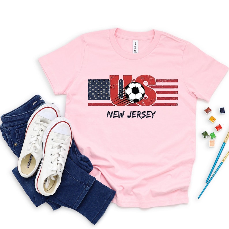 May include: A light pink short-sleeve t-shirt featuring a graphic of the American flag with the letters US, a soccer ball, and the text NEW JERSEY. The shirt is displayed with a pair of dark blue denim jeans and white canvas sneakers.