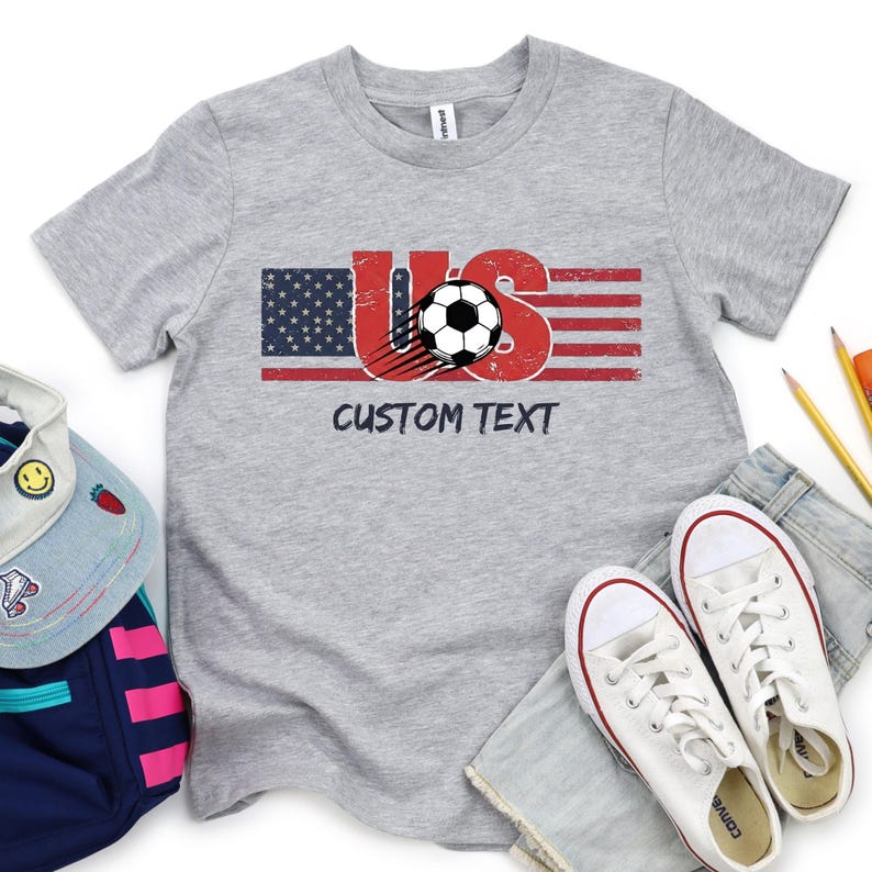 May include: A heather gray short-sleeve t-shirt featuring a graphic of the letters US with a distressed American flag pattern, a black and white soccer ball, and the text CUSTOM TEXT printed in black lettering below the graphic design.