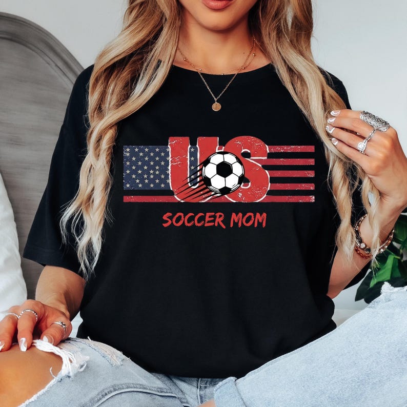 May include: A black t-shirt featuring a graphic design with the letters US, where the U is styled with a blue and white star pattern and the S is styled with red and white stripes. A black and white soccer ball is centered, with the text SOCCER MOM below.