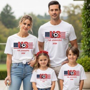 May include: A white short-sleeve t-shirt featuring a graphic of the American flag, a soccer ball, and the text US, THE JOHNSONS, and 2026. The design has a distressed, vintage-inspired texture with red, white, and blue colors throughout the print.