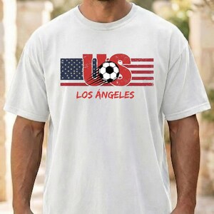 May include: A white short-sleeved t-shirt featuring a graphic of the American flag with the letters US in red, a black and white soccer ball, and the text LOS ANGELES printed in red underneath the flag design on the front of the garment.