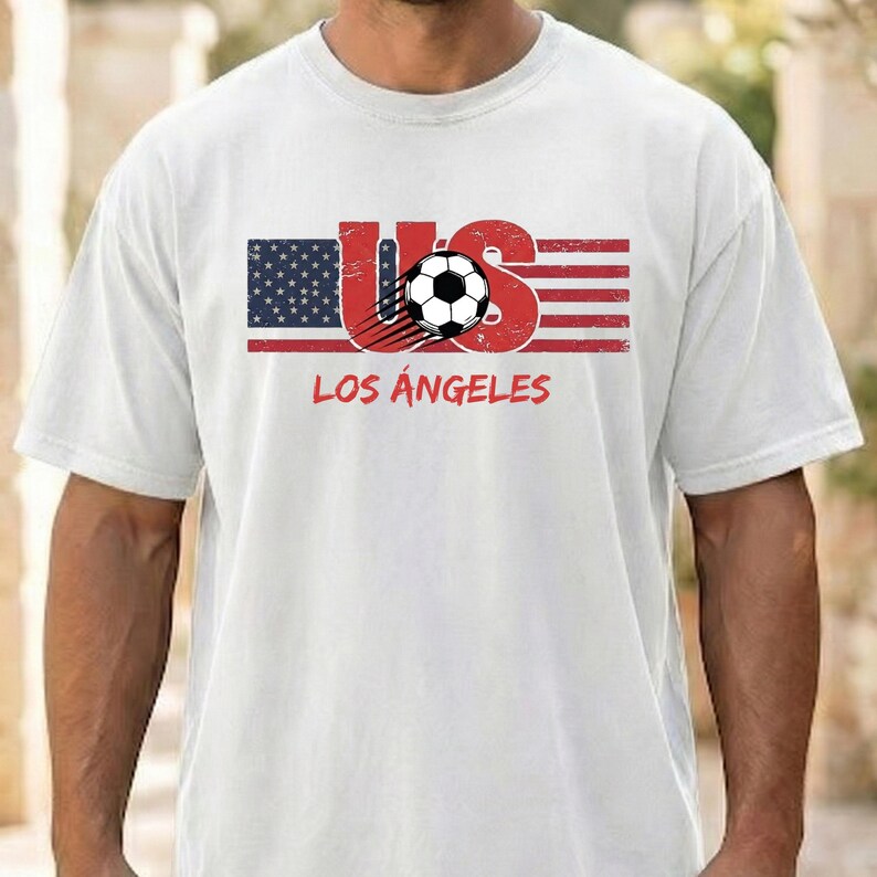 May include: A white short-sleeved t-shirt featuring a graphic of the American flag with the letters US in red, a black and white soccer ball, and the text LOS ANGELES printed in red underneath the flag design on the front of the garment.