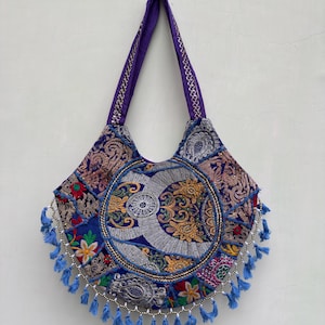 May include: A purple shoulder bag featuring a circular central panel with silver embroidery and intricate floral patterns. The bag is decorated with colorful fabric patches, metallic thread accents, and a row of light blue tassels along the bottom edge.