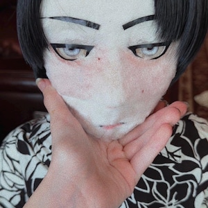 May include: A person wearing a mask with a pale face, black hair, and stylized eyes. The person is wearing a black and white floral print shirt.