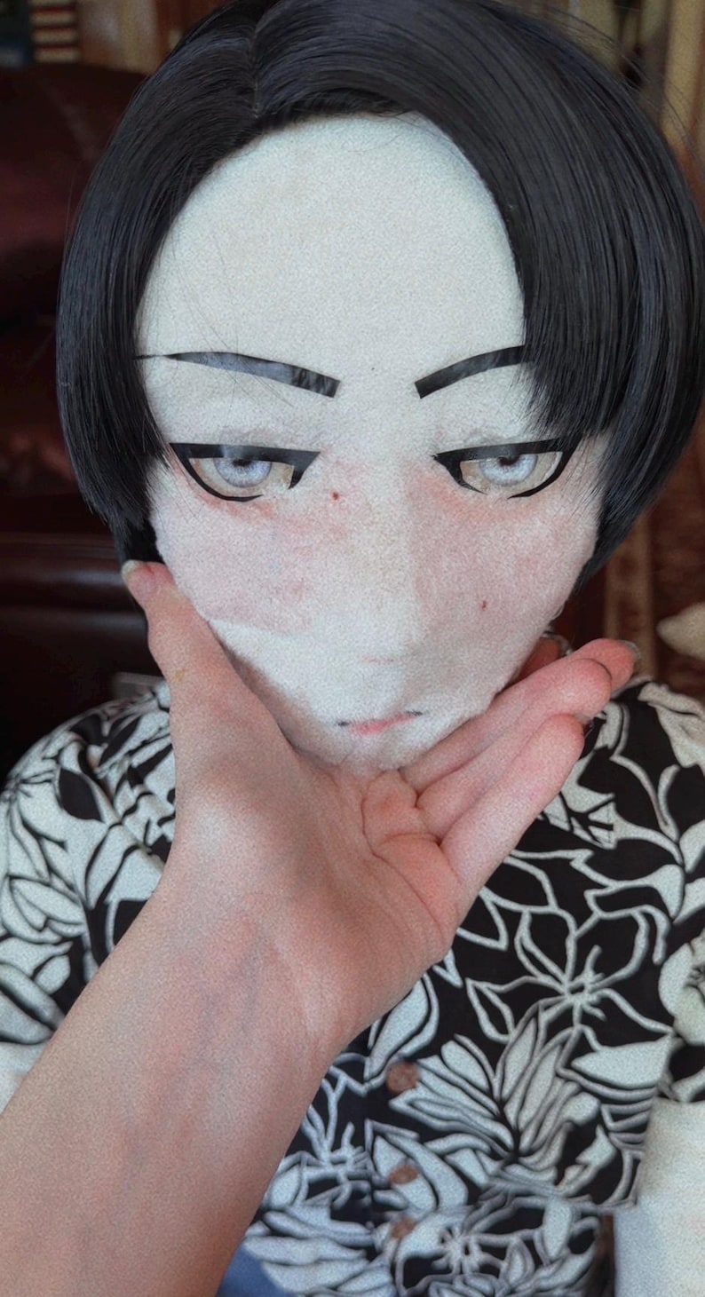 May include: A person wearing a mask with a pale face, black hair, and stylized eyes. The person is wearing a black and white floral print shirt.