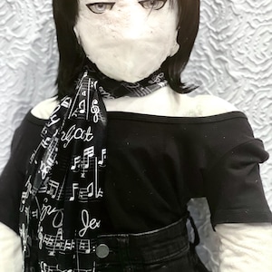 May include: A doll wearing a black off-the-shoulder top, black jeans, and a black and white scarf. The scarf is printed with musical notes and the words "Je dis it". The doll has black hair and a pale face.