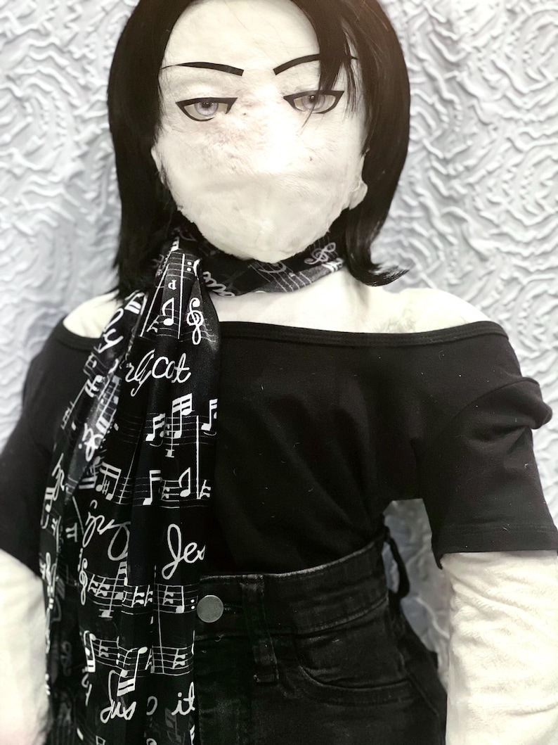 May include: A doll wearing a black off-the-shoulder top, black jeans, and a black and white scarf. The scarf is printed with musical notes and the words "Je dis it". The doll has black hair and a pale face.
