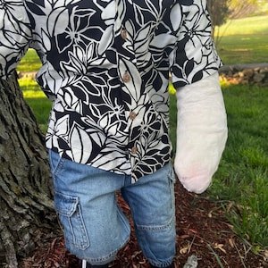 May include: A black and white floral print button-down shirt paired with denim cargo shorts. The outfit is completed with black sneakers. The shirt features a tropical leaf design.