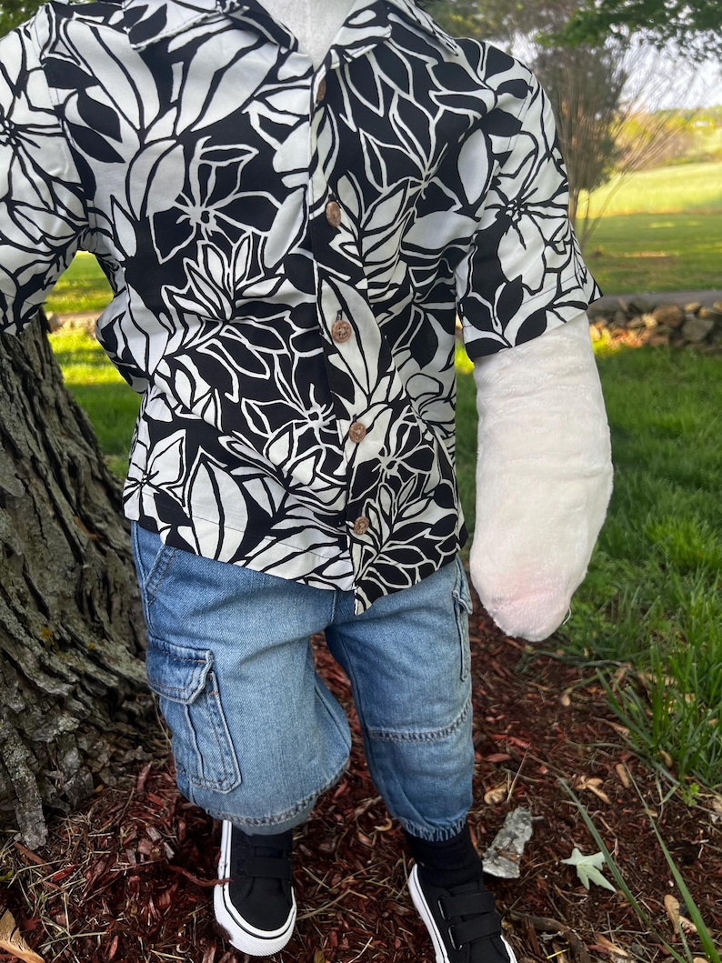 May include: A black and white floral print button-down shirt paired with denim cargo shorts. The outfit is completed with black sneakers. The shirt features a tropical leaf design.