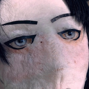 May include: Close-up of a plush doll face with black eyeliner and eyebrows. The doll has light blue eyes and black hair. The skin is a pale pink color.