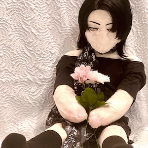 May include: A plush doll with black hair and a black outfit, holding pink and white flowers. The doll has a fabric face and is wearing a scarf with musical notes. The doll is sitting on a white, textured surface.