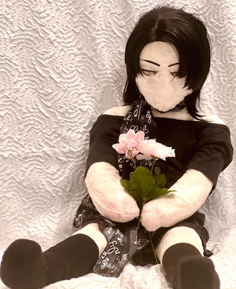 May include: A plush doll with black hair and a black outfit, holding pink and white flowers. The doll has a fabric face and is wearing a scarf with musical notes. The doll is sitting on a white, textured surface.