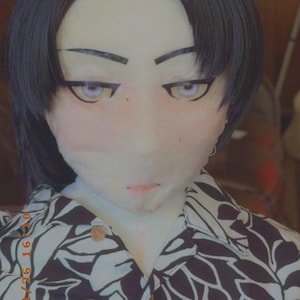 May include: A doll head with black hair and stylized facial features, including large eyes and a small mouth. The doll is wearing a button-down shirt with a black and white floral pattern.