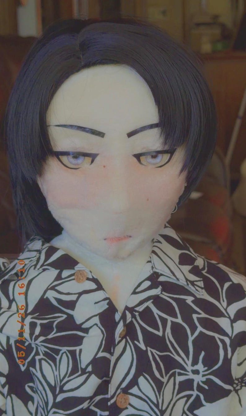 May include: A doll head with black hair and stylized facial features, including large eyes and a small mouth. The doll is wearing a button-down shirt with a black and white floral pattern.