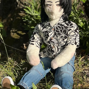 May include: A plush doll with black hair, wearing a black and white floral print shirt, blue jeans, and white sneakers with brown soles. The doll is seated in a grassy area, with sunlight filtering through.