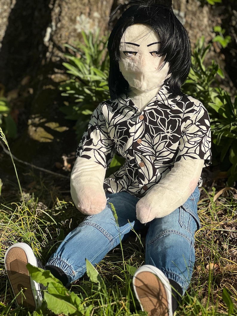 May include: A plush doll with black hair, wearing a black and white floral print shirt, blue jeans, and white sneakers with brown soles. The doll is seated in a grassy area, with sunlight filtering through.