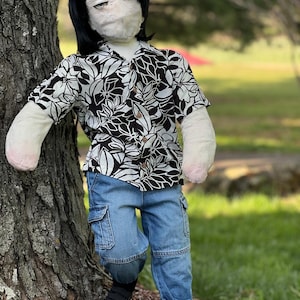 May include: A doll dressed in a black and white floral print button-down shirt and blue cargo pants. The doll has black hair and is wearing black and white sneakers. The doll is leaning against a tree in a grassy outdoor setting.