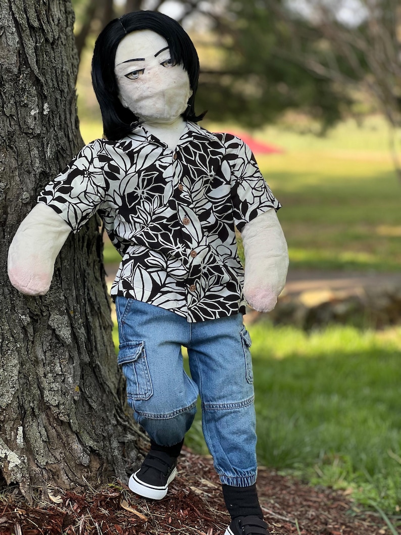 May include: A doll dressed in a black and white floral print button-down shirt and blue cargo pants. The doll has black hair and is wearing black and white sneakers. The doll is leaning against a tree in a grassy outdoor setting.