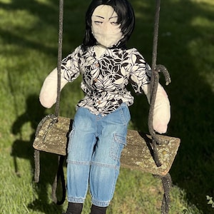 May include: A handmade doll with black hair, wearing a black and white floral shirt, blue jeans, black socks, and black sneakers. The doll is seated on a wooden swing, suspended by black ropes, against a backdrop of green grass and sunlight.