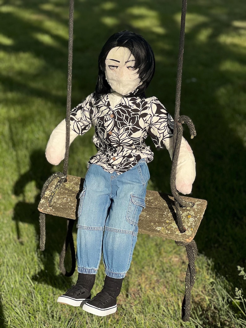 May include: A handmade doll with black hair, wearing a black and white floral shirt, blue jeans, black socks, and black sneakers. The doll is seated on a wooden swing, suspended by black ropes, against a backdrop of green grass and sunlight.