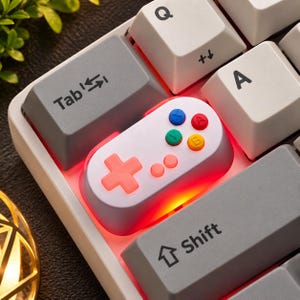May include: A mechanical keyboard keycap shaped like a retro video game controller, featuring a white base, a red directional pad, and four small colored action buttons in blue, red, green, and yellow, illuminated by a bright red backlight.