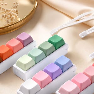 May include: Four white rectangular mechanical keyboard switch testers, each holding four colorful keycaps in various shades including brown, orange, pink, purple, green, light green, blue, and white, accompanied by two white lanyards.