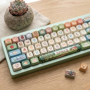 Könnte beinhalten: Eine pastellfarbene Tastatur mit individuellen Tastenkappen mit verschiedenen Illustrationen, darunter eine Katze, ein Apfel und Berge. Die Tastatur hat einen hellgrünen Rahmen und enthält eine "SPACE"-Taste mit einem Landschaftsdesign. Eine kleine Sukkulente in einem Steintopf befindet sich in der Nähe.