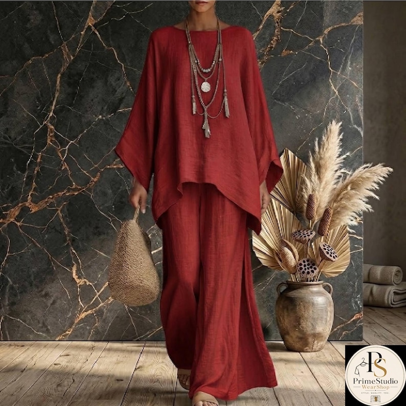 May include: A red linen two-piece outfit, featuring a long-sleeved top and wide-leg pants. The top has a unique asymmetrical hemline. A woven bag and layered necklace complete the look. A decorative vase with dried plants is in the background.
