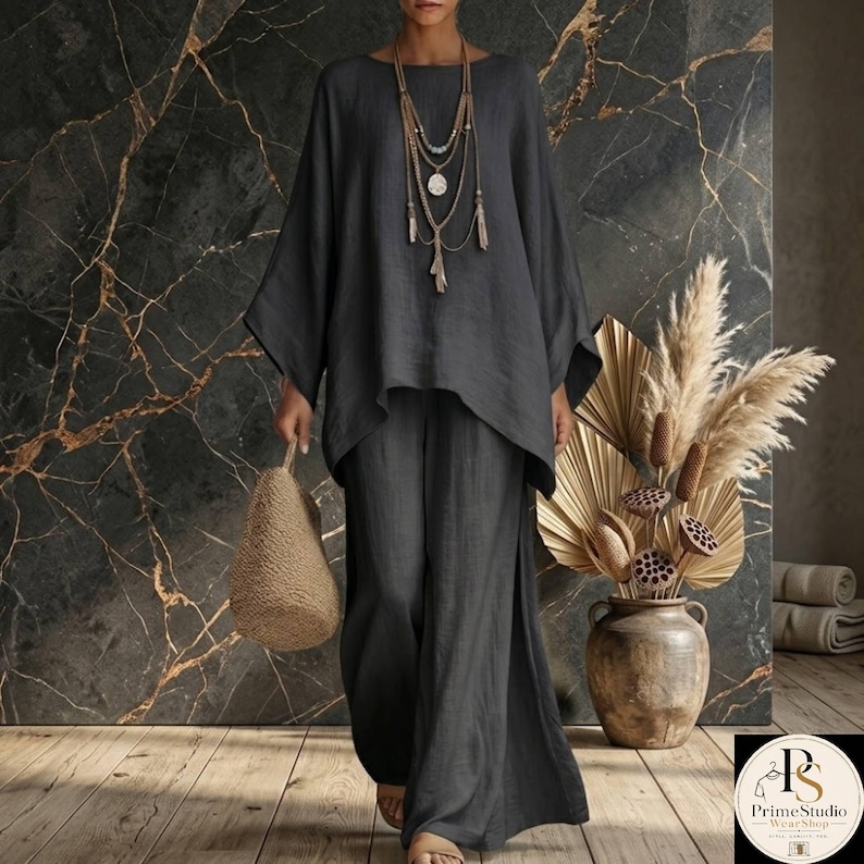 May include: A charcoal gray linen two-piece outfit, featuring a loose-fitting top with wide sleeves and matching wide-leg pants. The outfit is accessorized with layered necklaces and a woven handbag. The background includes a marble wall and a vase of dried plants.