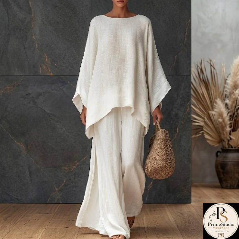 May include: A two-piece white linen outfit. The top has a wide, asymmetrical hem and long sleeves. The pants are wide-legged with a slit on the side. A woven tote bag is held in the hand. The outfit is suitable for casual wear.
