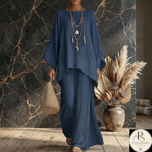 May include: A two-piece navy blue linen outfit. The top has a round neck and long, wide sleeves. The pants are wide-legged with a side slit. A woven bag and layered necklace complete the look. The background is a dark marble wall.