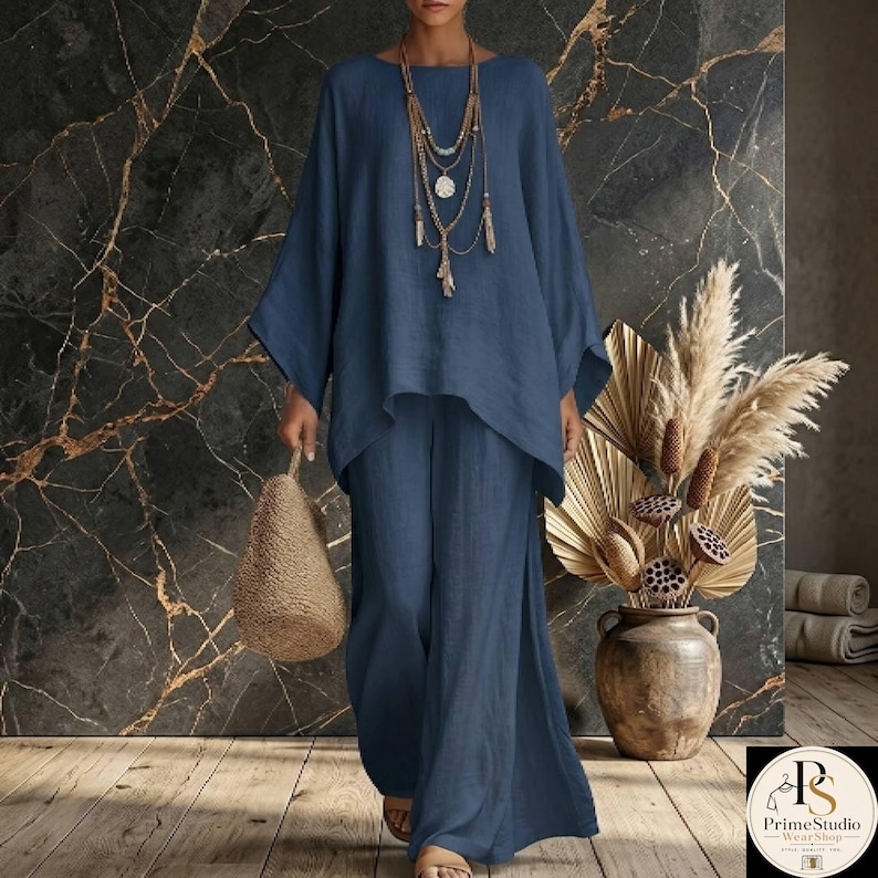 May include: A two-piece navy blue linen outfit. The top has a round neck and long, wide sleeves. The pants are wide-legged with a side slit. A woven bag and layered necklace complete the look. The background is a dark marble wall.