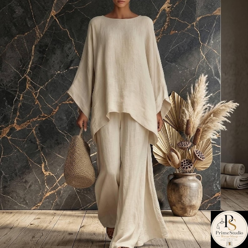 May include: A beige linen two-piece outfit, featuring a long-sleeved top with a wide neckline and matching wide-leg pants. The outfit is paired with a woven tote bag. The background has a dark marble effect.