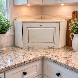 May include: A corner cabinet with a distressed, off-white finish. The cabinet has a hinged door with a central panel and metal hardware. It sits on a granite countertop in a kitchen setting. A wooden cutting board and a white pitcher are visible.