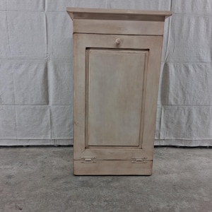 May include: A tall, rectangular wooden cabinet finished in a distressed off-white paint. The cabinet features a single vertical door with a small central knob, a flat top surface, and two visible metal hinges at the bottom of the front panel.