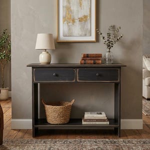 May include: A black wooden console table with two drawers featuring distressed edges and round knobs. The table has a lower shelf holding a woven basket and a stack of books. The top surface holds a beige table lamp and three stacked books.