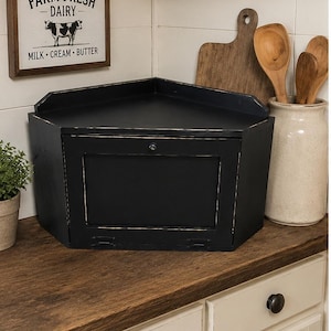 May include: A black, corner-shaped bread box with a single door and a small lock. The box sits on a wooden countertop, with a framed sign that reads "Farm Fresh Dairy" above it. A small plant and a ceramic jar with wooden spoons are also visible.