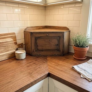May include: A wooden corner bread box with a hinged front door and a small metal knob. The bread box features a dark brown finish and is positioned on a wooden countertop in a kitchen corner, next to a wooden cutting board and a potted plant.