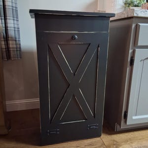 May include: A black wooden trash can with a decorative X design on the front. The can has a small knob and hinges. The trim has a distressed, off-white finish. The trash can is rectangular and stands upright.