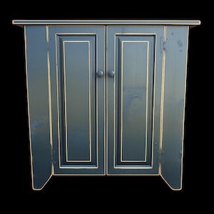 May include: A dark blue cabinet with two doors and a distressed finish. The cabinet has a rectangular shape with a slightly overhanging top and a decorative trim. The doors have round knobs and are outlined with a gold trim.