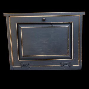 May include: A black, wall-mounted cabinet with a single door. The cabinet has a distressed finish with gold trim and hardware. The door is rectangular with a recessed panel and a small, round knob. Hinges are visible at the bottom.