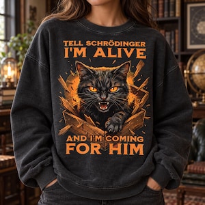 Op de afbeelding: Een antracietgrijze sweater met ronde hals en een afbeelding van een zwarte kat met gloeiende oranje ogen die uit een houten kist komt. De tekst "TELL SCHRÖDINGER I'M ALIVE AND I'M COMING FOR HIM" staat in oranje letters boven en onder de kat.