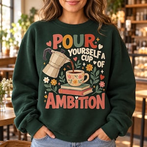 Puede incluir: Una sudadera de cuello redondo verde oscuro con un estampado gráfico de una cafetera italiana vertiendo café en una taza floral sobre dos libros. El texto "POUR YOURSELF A CUP OF AMBITION" aparece en letras coloridas de estilo retro con corazones y flores.