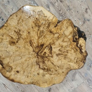 May include: A round, live-edge wooden table top featuring a natural light tan and dark brown wood grain pattern. The surface has a glossy, smooth finish that highlights the intricate, organic cracks and textures of the raw wood slab.