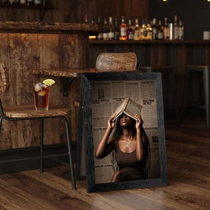 May include: A framed vertical print featuring a person with long dark braids wearing a brown tank top, holding an open book over their face. The background consists of a collage of newspaper clippings with text including Voices from the Past.
